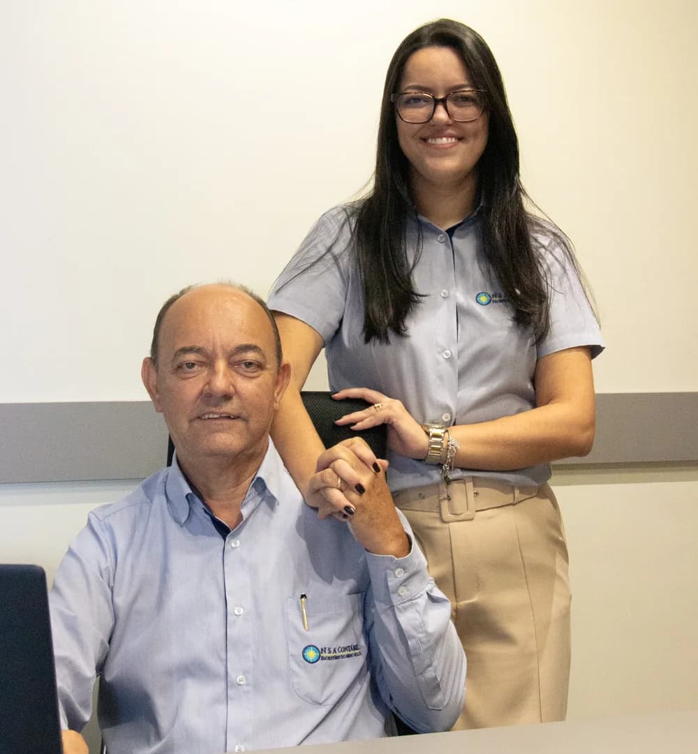 José, fundador do escritório, sentado à mesa, e Letícia, sucessora e gestora, em pé ao lado dele