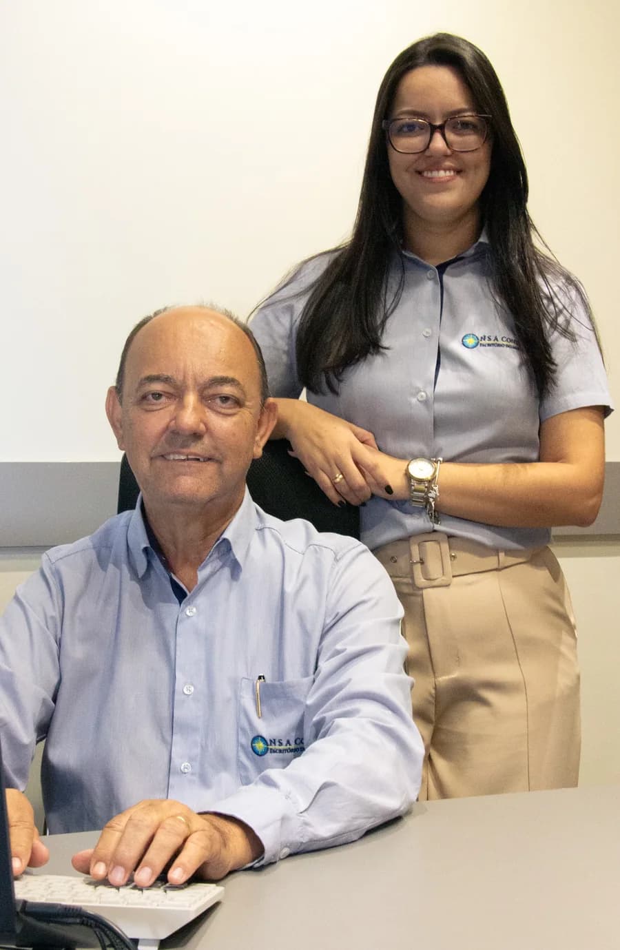 José, fundador do escritório, sorrindo à sua mesa de trabalho, com Letícia, sucessora e gestora, em pé atrás dele